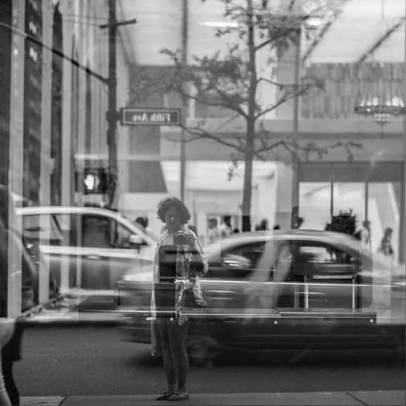 Black and white street frame: a woman stands at the kerb on Fifth Avenue, traffic and shopfronts doubled by a reflection across the whole image.